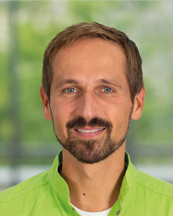 Portraitfoto von Zahnarzt Dr. Wenninger, freundlich lächelnd in grüner Arbeitskleidung.